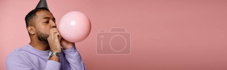 Photo for Young African American man with braces happily blows up a balloon while wearing a party hat on a pink background. - Royalty Free Image