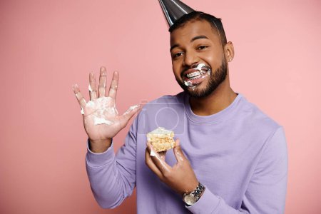 Young African American man in party hat happily eating a birthday cake on a pink background.
