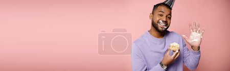 Photo for A happy, young African American man with braces, in a purple shirt, savoring a pastry on a pink background. - Royalty Free Image