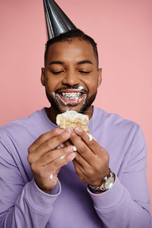 Junger Afroamerikaner in Hosenträgern isst fröhlich ein Sandwich, während er einen Partyhut auf rosa Hintergrund trägt.