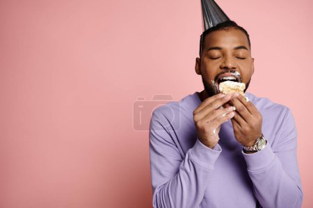 Joven, alegre hombre afroamericano con frenos come alegremente un pastel de cumpleaños mientras usa un sombrero de fiesta sobre un fondo rosa.