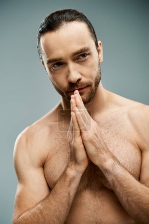 A handsome, shirtless man with a beard holds his hands together in a serene and strong gesture on a grey studio background.