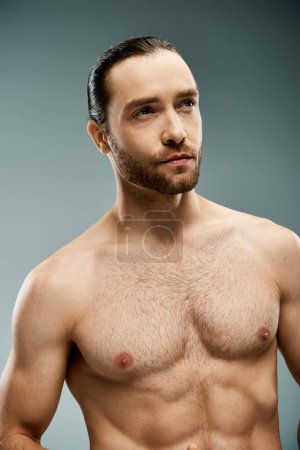 A handsome shirtless man with a beard striking a pose in a studio against a grey background.