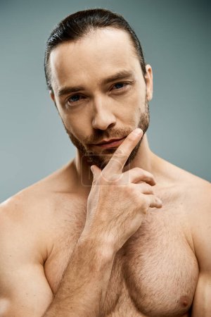 A handsome shirtless man with a beard striking a confident pose against a grey studio backdrop.