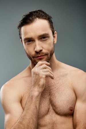 A shirtless man with a beard, deep in thought, hand resting on his chin, against a grey studio backdrop.