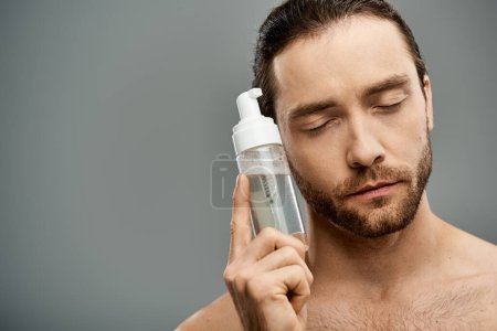 Téléchargez les photos : Un homme torse nu et barbu tient avec confiance une bouteille de nettoyant sur fond de studio gris. - en image libre de droit