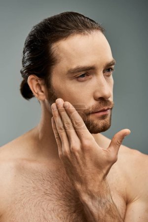 Handsome shirtless man with a beard, deep in thought, resting his hand on his face against a grey studio backdrop.