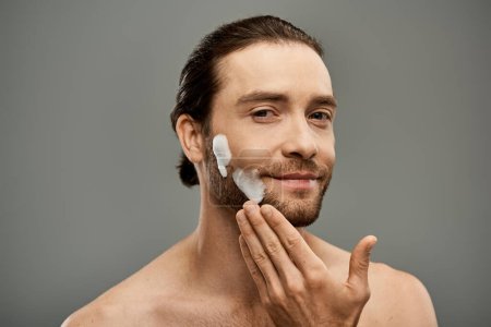 Foto de Un hombre sin camisa con barba usa espuma en su cara en un estudio sobre un fondo gris. - Imagen libre de derechos