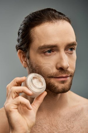Un hombre sin camisa con barba sosteniendo delicadamente un frasco de crema y aplicándolo en la cara contra un fondo gris de estudio.
