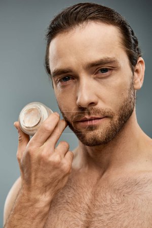 Un hombre sin camisa con barba sostiene delicadamente un frasco de crema en un ambiente de estudio con un fondo gris.