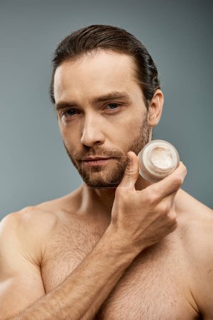 Foto de Hombre guapo y sin camisa con barba sosteniendo un frasco de crema en un estudio sobre un fondo gris. - Imagen libre de derechos