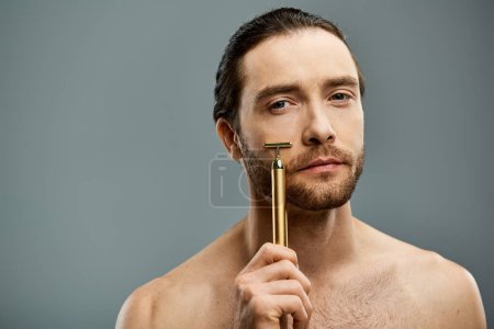 Shirtless man with beard holding golden razor in front of face against grey background.