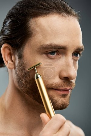 Shirtless bearded man carefully holds a golden razor in his hand against a grey studio backdrop.