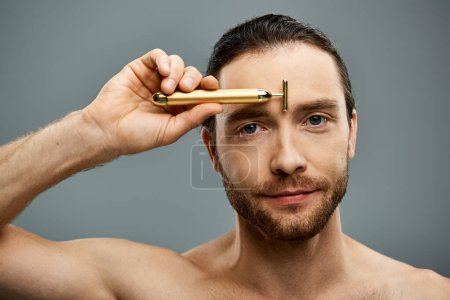 Shirtless man with beard touching golden razor to his forehead on a grey studio background.