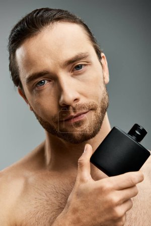 Téléchargez les photos : Un bel homme torse nu avec une barbe tient une bouteille de parfum sur un fond gris dans un décor de studio. - en image libre de droit