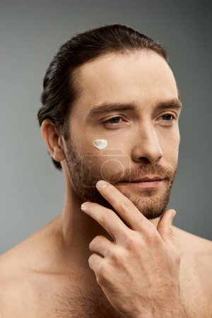 Foto de Un hombre guapo y sin camisa con barba se pone una máscara de crema en la cara en un estudio. - Imagen libre de derechos