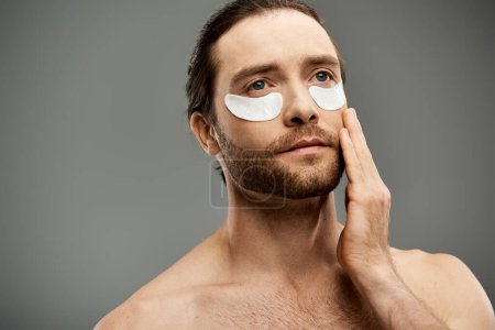 A shirtless, handsome man with a beard wearing eye patches looks intense on a grey studio background.
