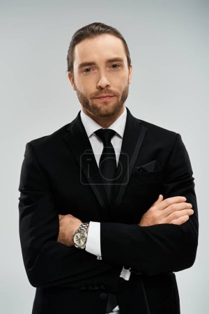 Un hombre de negocios barbudo con un traje elegante y corbata cruza con confianza sus brazos contra un fondo gris del estudio.