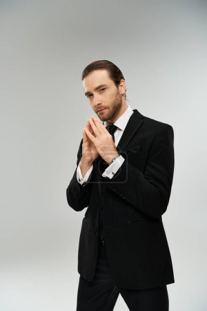 A bearded businessman in a suit and tie stands with hands clasped together in a thoughtful pose against a grey background.