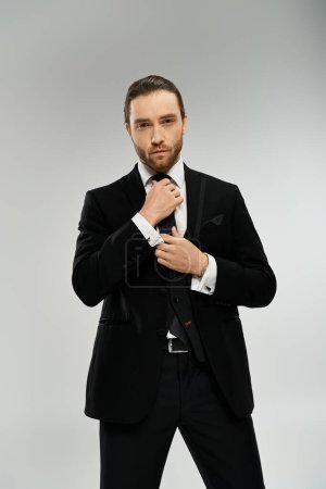 Bearded businessman in a suit adjusting his tie against a grey studio background.