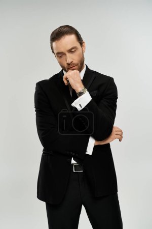 Un hombre de negocios guapo y barbudo con esmoquin posa con confianza para la cámara en un estudio sobre un fondo gris.