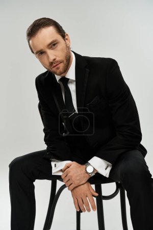 A handsome, bearded businessman in a stylish suit sits thoughtfully on a chair in a studio with a grey background.