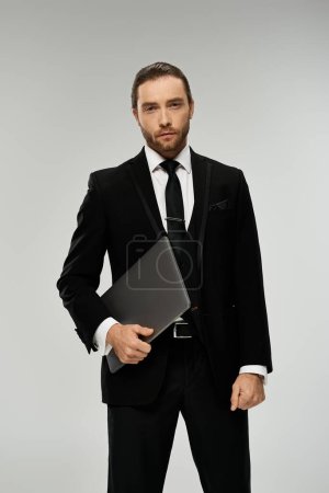 A handsome, bearded businessman in a suit confidently holds a laptop in a studio set against a grey background.