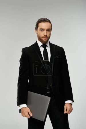 A bearded businessman in a sharp suit confidently holds a laptop in a sleek, professional studio setting.