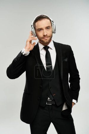 Foto de Un hombre de negocios barbudo en un elegante traje disfrutando de la música con auriculares en contra de un fondo gris estudio. - Imagen libre de derechos