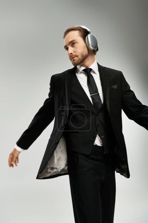 Foto de Un guapo hombre de negocios barbudo con un traje escuchando música con auriculares en un estudio sobre un fondo gris. - Imagen libre de derechos
