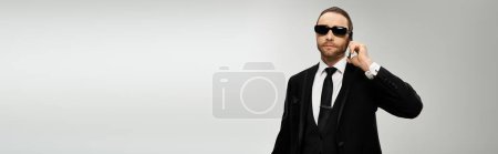 Bearded businessman in a suit and sunglasses talking on a cell phone against a grey studio background.