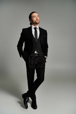 Ein gutaussehender und bärtiger Geschäftsmann in Anzug und Krawatte posiert selbstbewusst vor grauem Studiohintergrund.