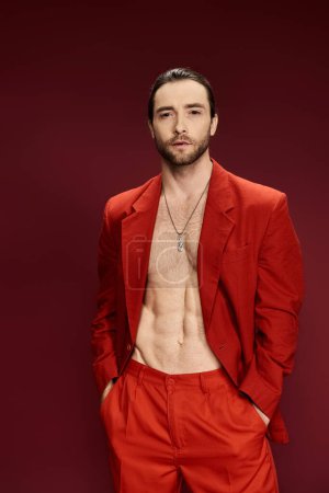 A handsome man in a striking red suit, sans shirt, confidently poses in a studio setting.
