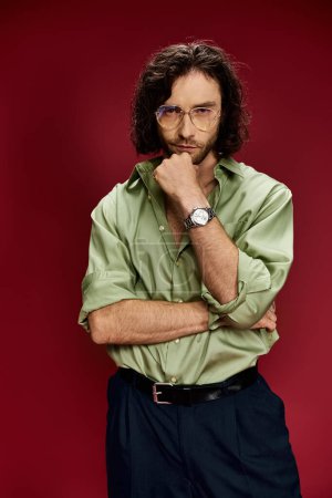A handsome man in glasses and a green silk shirt showcases a luxurious watch on his left hand against a bold red background.
