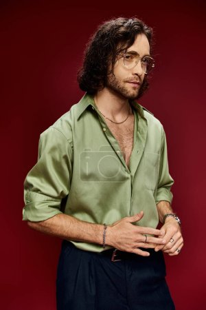 Foto de A handsome man in glasses strikes a pose in a green silk shirt and black pants against a vibrant red backdrop. - Imagen libre de derechos