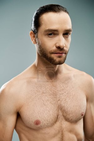 A handsome man with a beard stands shirtless against a grey backdrop in a studio setting.