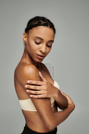 Young African American woman in strapless top with arm around her shoulder, showing self-love on grey background.