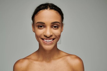 Beautiful young African American woman in a strapless top, exuding joy as she cares for her skin, set against a grey background.