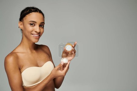 Young African American woman in a bra taking care of her skin, holding a container of cream on a grey background.