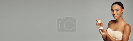 Photo for Young African American woman in a bra delicately holding a white cream while caring for her skin on a grey background. - Royalty Free Image