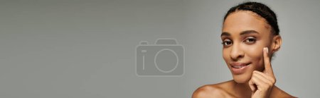 Foto de Joven mujer afroamericana en top sin tirantes tocándose delicadamente la cara, irradiando belleza y serenidad sobre fondo gris. - Imagen libre de derechos