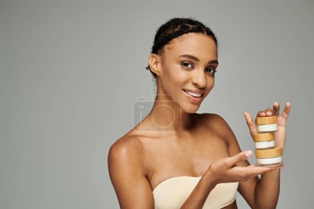 Foto de A young African American woman holding cream jars on a grey background. - Imagen libre de derechos