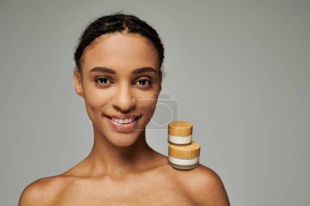 African American woman in strapless top smiles, beauty jars with cream on grey background.