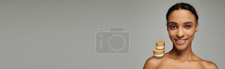 African American woman in strapless top smiles, beauty jars with cream on grey background.
