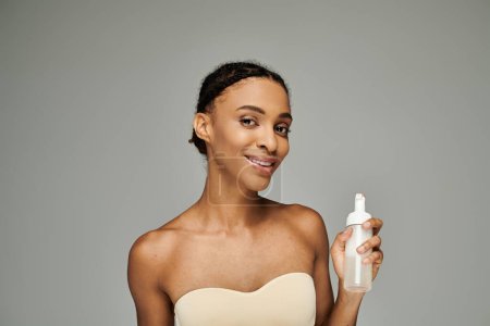Eine junge Afroamerikanerin mit makelloser Haut hält eine Flasche Gesichtsreiniger anmutig und ausbalanciert vor grauem Hintergrund.