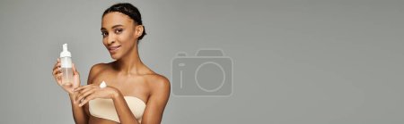 Foto de Young African American woman in a bikini holding a bottle of foam cleanser against a grey background. - Imagen libre de derechos