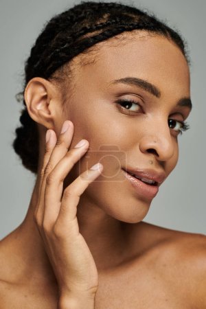 Young African American woman with braids posing confidently, caring for her skin on a grey background.