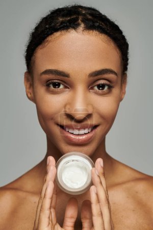 Photo for African American woman holds cream jar in front of face against grey background, taking skincare seriously. - Royalty Free Image