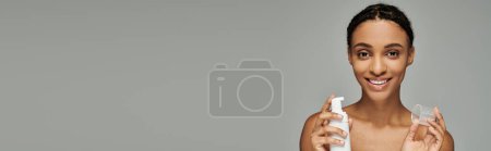 Photo for Young African American woman in strapless top nourishing her skin with a bottle of cleanser on grey background. - Royalty Free Image