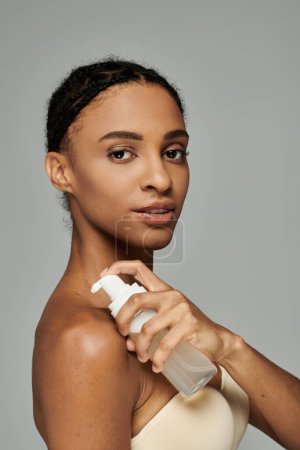 Foto de Young African American woman holding bottle of cleanser to moisturize skin, wearing strapless top on grey background. - Imagen libre de derechos